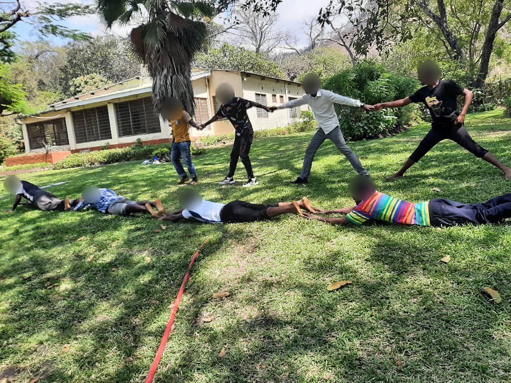 Eine Gruppe bildet beim Teambuilding eine Kette über eine Wiese, im Hintergrund ein Gebäude